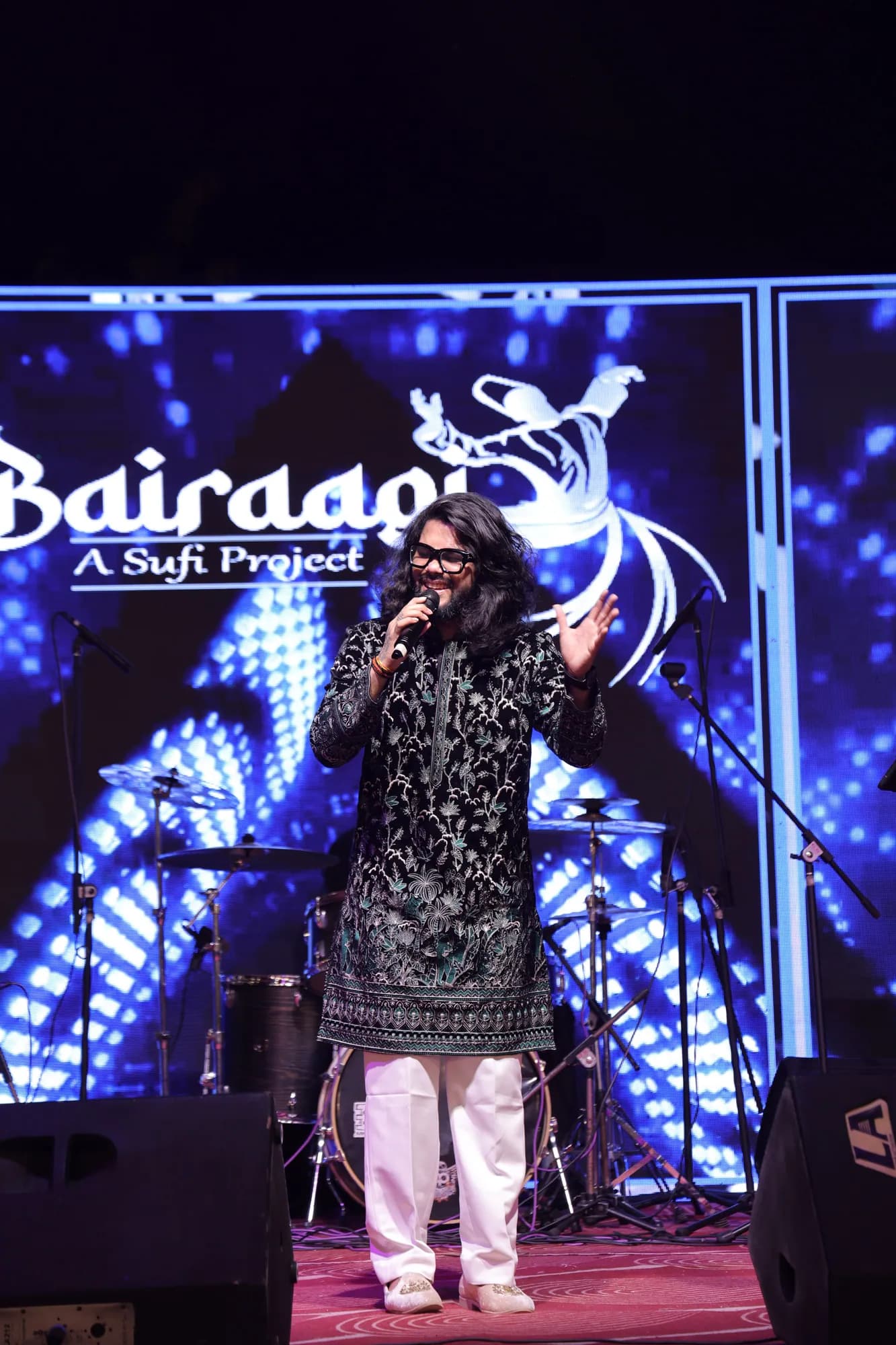 Softrock's vocalist in a black embroidered kurta singing in front of a blue LED screen reading Bairagi, A Sufi Project.
