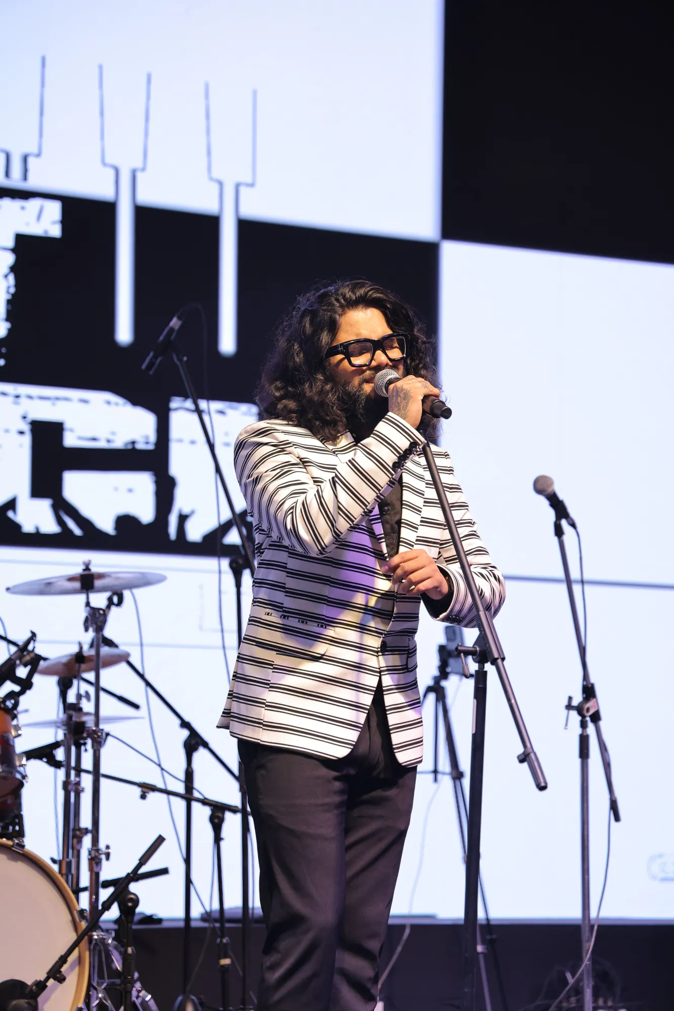 Softrock's vocalist in a striped blazer singing in front of a bright stage screen.