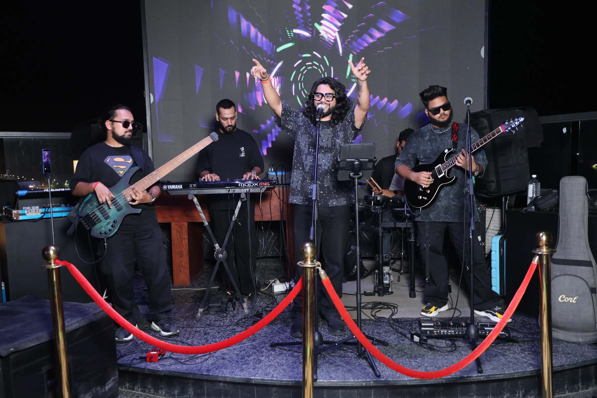 Softrock playing on a round stage behind red velvet ropes, the vocalist's arms raised between the bassist, keyboardist and guitarist.