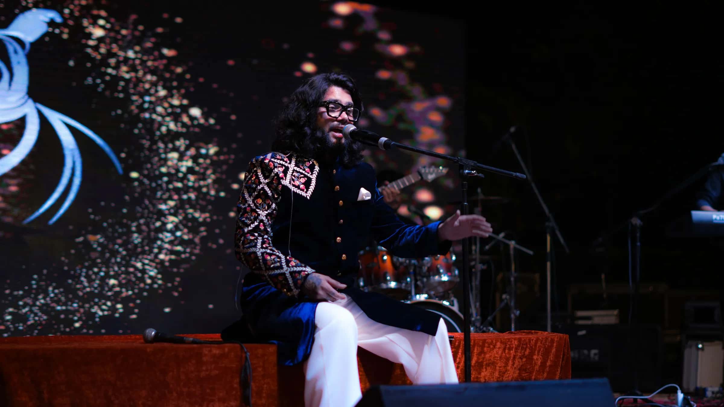 Softrock's vocalist seated on a low stage in an embroidered velvet jacket, singing against a glittering LED backdrop.